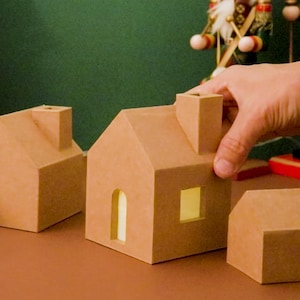 Könnte beinhalten: Karton-Weihnachtsdorfhäuser und eine kleine Schachtel sind auf einer braunen Oberfläche angeordnet. Eine Hand greift nach einem der Häuser. Ein kleiner Weihnachtsbaum mit roten Ornamenten und eine Schneekugel befinden sich im Hintergrund.