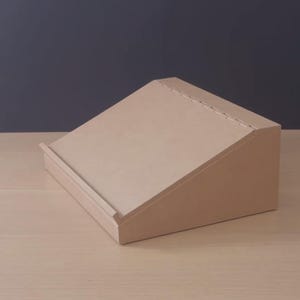 May include: A light brown, wedge-shaped wooden display stand with a smooth, flat angled surface and a small horizontal ledge at the bottom edge to support items. The stand features a simple, solid construction with clean lines and a matte finish.