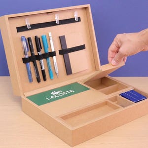 May include: A light brown wooden desk organizer with multiple compartments and a hinged lid. The organizer contains pens, a notepad, and a green rectangle with the Lacoste logo. A hand is opening the lid.