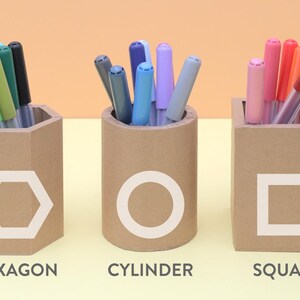 May include: Three cardboard pen holders in different shapes: hexagon, cylinder, and square. Each holder has a white geometric shape on the front and is filled with colorful markers. The text "CARDBOARD DIY" is on the left side of the image.