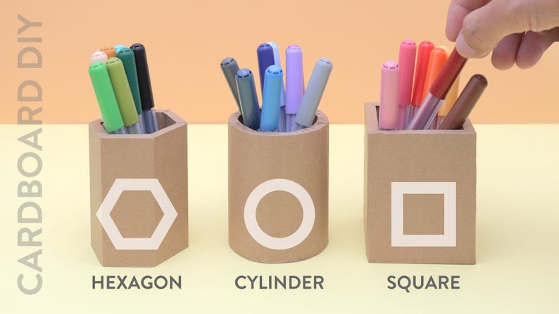 May include: Three cardboard pen holders in different shapes: hexagon, cylinder, and square. Each holder has a white geometric shape on the front and is filled with colorful markers. The text "CARDBOARD DIY" is on the left side of the image.