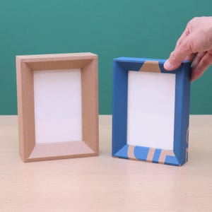 May include: Two rectangular picture frames. One is light brown, the other is blue with a brown accent. The frames have a white interior and are made of a cardboard-like material. The background is a solid green colour.