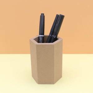 May include: A brown, hexagonal pen holder made of cardboard, containing three black pens. The pens are of varying sizes and styles. The background is a gradient of orange and yellow.