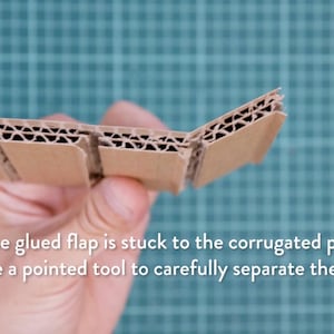 May include: A section of corrugated cardboard held in hand, showing the layered construction. The text reads: "If the glued flap is stuck to the corrugated part, use a pointed tool to carefully separate them."