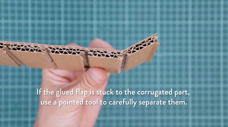 May include: A section of corrugated cardboard held in hand, showing the layered construction. The text reads: "If the glued flap is stuck to the corrugated part, use a pointed tool to carefully separate them."