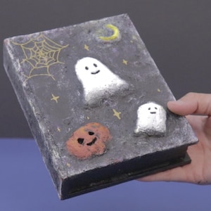 May include: A Halloween-themed book with a dark gray cover featuring a gold spiderweb, a yellow crescent moon, and two white ghosts. An orange pumpkin with a smiling face is also on the cover.
