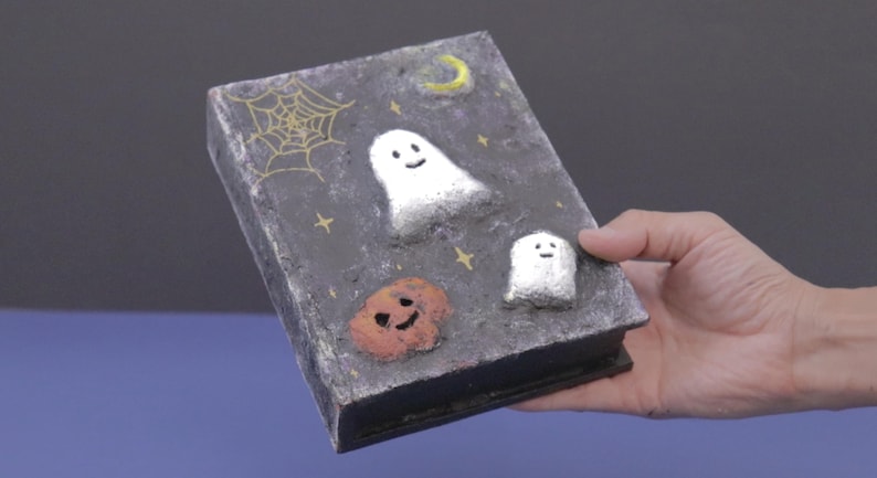 May include: A Halloween-themed book with a dark gray cover featuring a gold spiderweb, a yellow crescent moon, and two white ghosts. An orange pumpkin with a smiling face is also on the cover.