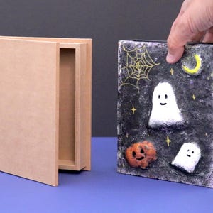 May include: A Halloween-themed box with a decorative lid featuring a night sky design. The lid has a spiderweb, moon, stars, and smiling ghosts and a pumpkin. The box is held by a hand and is next to an open, plain wooden box.