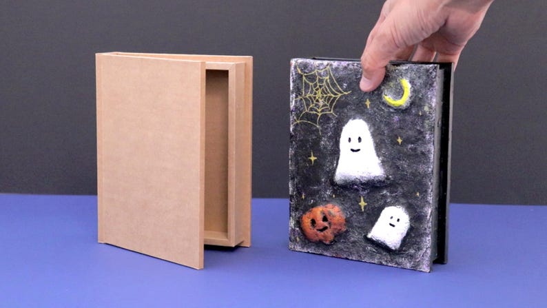 May include: A Halloween-themed box with a decorative lid featuring a night sky design. The lid has a spiderweb, moon, stars, and smiling ghosts and a pumpkin. The box is held by a hand and is next to an open, plain wooden box.