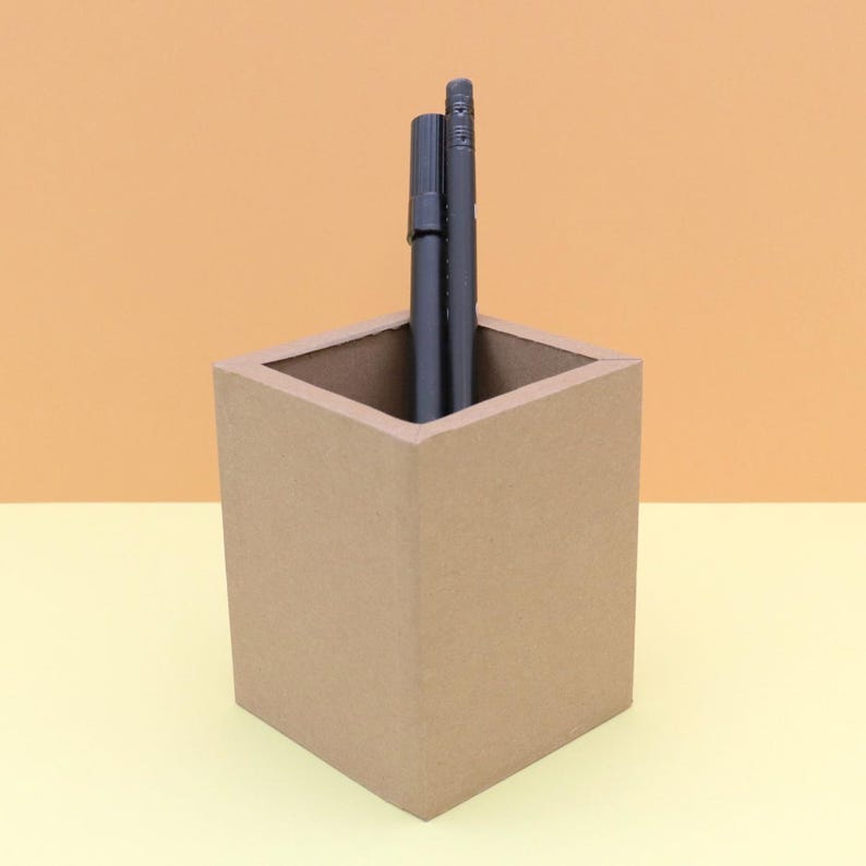 May include: A square, light brown pen holder with two black pens inside. The pen holder is made of a cardboard-like material and sits on a yellow surface with an orange background.