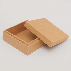 Puede incluir: Una caja rectangular de cartón abierta con una tapa separada. La caja y la tapa son de color marrón claro. La tapa está ligeramente entreabierta, revelando el interior de la caja. La caja está sobre una superficie blanca.