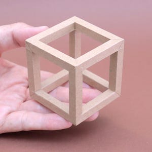 May include: A light brown, open-frame cube held in a hand. The cube is constructed of thin, rectangular pieces of material, creating the illusion of a three-dimensional shape. The background is a solid, muted brown.