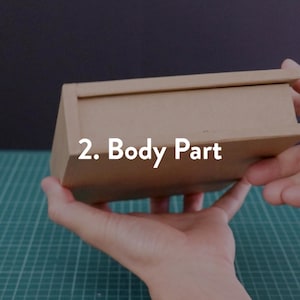 May include: A small, rectangular cardboard box with a lid, held in hands. The box is a light brown color. Text on the box reads "2. Body Part". The background is a dark color and a green cutting mat.
