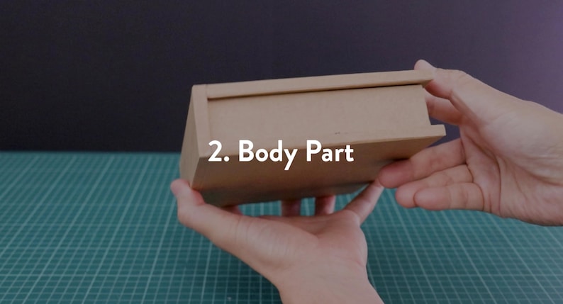 May include: A small, rectangular cardboard box with a lid, held in hands. The box is a light brown color. Text on the box reads "2. Body Part". The background is a dark color and a green cutting mat.