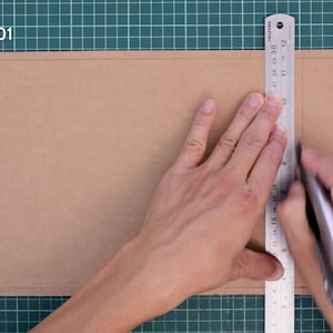 May include: A craft project in progress, showing a person using a metal ruler and a utility knife to cut a piece of brown cardboard. The text "Key technique 01 Half-Cut" is visible in the upper left corner. The cutting surface is a green grid.