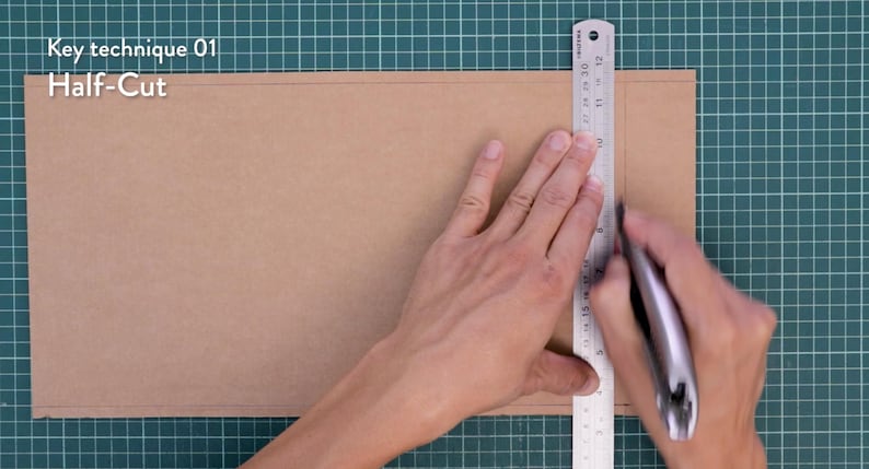May include: A craft project in progress, showing a person using a metal ruler and a utility knife to cut a piece of brown cardboard. The text "Key technique 01 Half-Cut" is visible in the upper left corner. The cutting surface is a green grid.