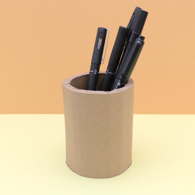 May include: A cylindrical, brown cardboard pen holder filled with several black pens. The pens are of varying thicknesses and have silver clips. The background is split between a light yellow and a light orange.
