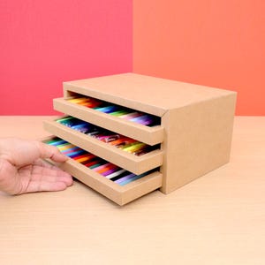 Puede incluir: Un organizador de tres cajones hecho de cartón, con cada cajón lleno de bolígrafos de colores. El cajón superior está cerrado, mientras que los dos inferiores están parcialmente abiertos, revelando los bolígrafos. El fondo es un degradado de rojo y naranja.