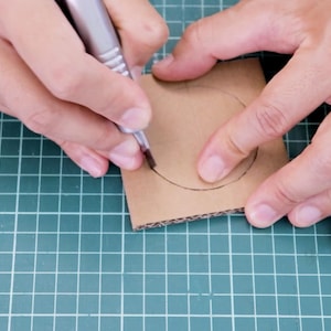 May include: A person is using a pen to draw a circle on a piece of brown cardboard. The cardboard is on a green grid surface. The pen is silver and black.