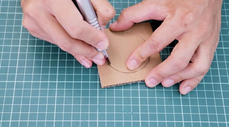 May include: A person is using a pen to draw a circle on a piece of brown cardboard. The cardboard is on a green grid surface. The pen is silver and black.