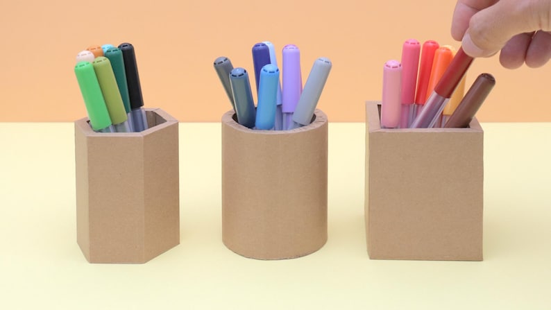 May include: Three brown pen holders with colorful markers. The holders are a hexagon, a cylinder, and a square. A hand is reaching into the square holder to grab a marker. The markers are various colors.