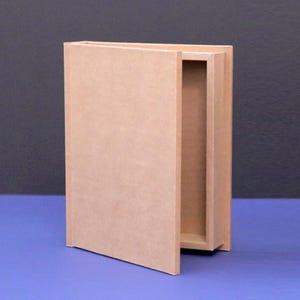 May include: A light brown, rectangular box designed to resemble a book. The box has a hinged front cover that is slightly ajar, revealing an empty interior. The box is made of a light brown material and sits on a blue surface.