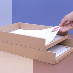 Pode incluir: Um organizador de mesa de escritório de cartão castanho claro de dois níveis, com uma mão a alcançar um documento. O organizador contém papéis brancos e está sobre um fundo de dois tons castanho e azul.
