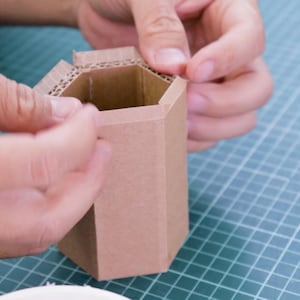 May include: A person is assembling a small, hexagonal cardboard container. The container is made of brown corrugated cardboard. The person's hands are visible as they fold and secure the top flaps of the container.