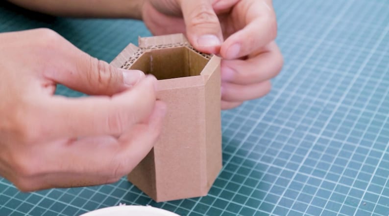 May include: A person is assembling a small, hexagonal cardboard container. The container is made of brown corrugated cardboard. The person's hands are visible as they fold and secure the top flaps of the container.
