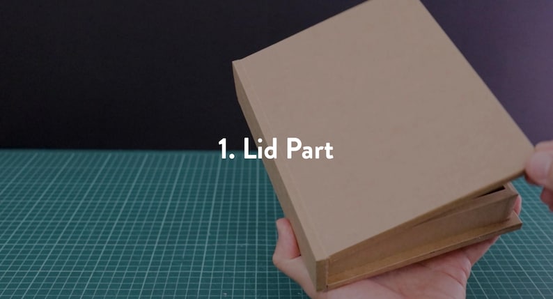 May include: A partially opened, rectangular cardboard box with the text "1. Lid Part" in white. The box is held by a hand, revealing the interior. The background is a mix of black and green.