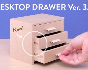 May include: A small, three-drawer desktop organiser made of light brown cardboard. The text "DESKTOP DRAWER Ver. 3.0" is visible at the top. The word "New!" is also visible. A hand is pulling open one of the drawers.