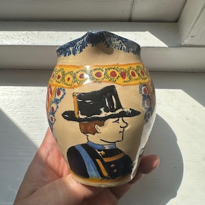 Puede incluir: Jarra de cerámica pintada a mano con un retrato de un hombre con sombrero negro y uniforme. La jarra tiene una base beige con una banda floral amarilla y roja alrededor del centro y detalles azules en el borde.
