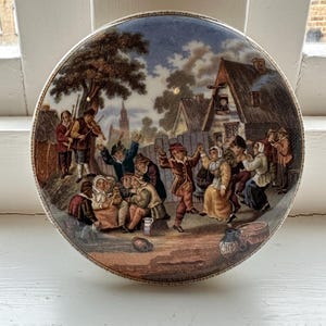 Op de afbeelding: Een rond keramiek deksel met een kleurrijke scène van mensen die dansen en muziek maken in een dorp. Het deksel is versierd met bruine, blauwe en witte verf en heeft een tekstinscriptie met de tekst "The Village Wedding".