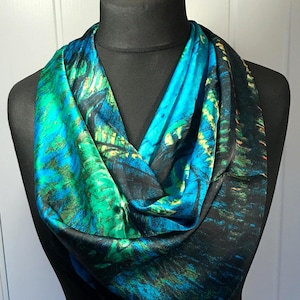 Pode incluir: Um lenço vibrante e estampado em tons de verde-azulado, turquesa e preto, drapeado sobre um manequim preto. O design do lenço apresenta uma mistura de cores, criando um efeito artístico e fluido. O lenço é de seda.