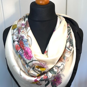 May include: A cream silk scarf with a colourful floral design, including pink, yellow, and blue flowers. The scarf has a black border and is displayed on a black mannequin head.