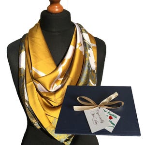 May include: A gold silk scarf with a floral pattern and a white background. The scarf is folded and displayed on a mannequin. A blue gift box with a gold ribbon and a gift tag that says "Especially for you" and "Our Auwie Crown" is in front of the scarf.