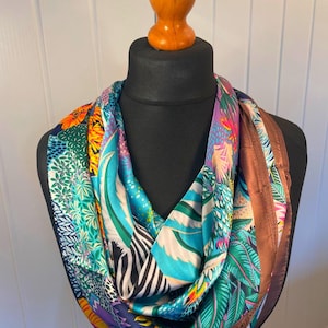 May include: A colorful silk scarf with a vibrant print of tropical leaves, zebra stripes, and floral patterns. The scarf features a mix of blues, greens, oranges, and purples, with a brown border. It is displayed on a black mannequin.