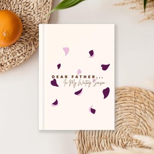 May include: A cream-coloured journal with the text "DEAR FATHER... In My Waiting Season" and purple petal designs. The journal is on a white surface with oranges and a woven basket in the background.