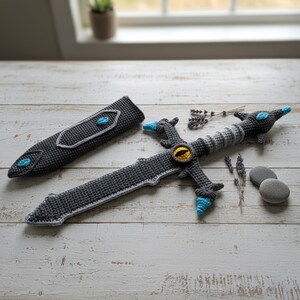 May include: A crocheted sword and sheath set in shades of gray, with blue and yellow accents. The sword features a detailed hilt with an eye design and a textured blade. The sheath has a matching design. A unique handmade item.