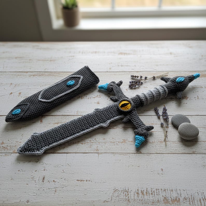 May include: A crocheted sword and sheath set in shades of gray, with blue and yellow accents. The sword features a detailed hilt with an eye design and a textured blade. The sheath has a matching design. A unique handmade item.