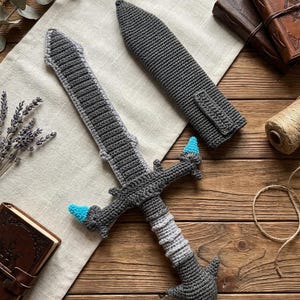 May include: A crocheted sword and sheath set in shades of gray and light blue. The sword features a detailed handle and blade, with blue accents on the hilt. The sheath is a matching gray, designed to fit the sword. The set is displayed on a wooden surface.