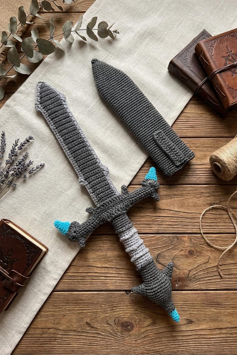 May include: A crocheted sword and sheath set in shades of gray and light blue. The sword features a detailed handle and blade, with blue accents on the hilt. The sheath is a matching gray, designed to fit the sword. The set is displayed on a wooden surface.
