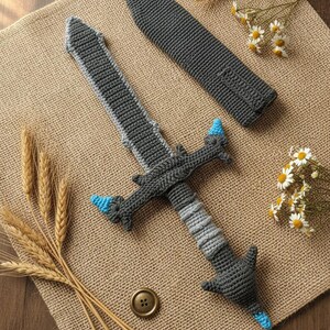 May include: A handmade crochet sword with a matching sheath. The sword features a gray blade with light gray and blue accents. The handle is gray and light gray. The sheath is gray. The items are displayed on a burlap surface.