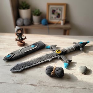 May include: Crocheted gray sword with blue and yellow accents, a matching sheath, and a small figurine. The sword features a detailed handle and blade design. A ball of gray yarn and a small stone are also present.