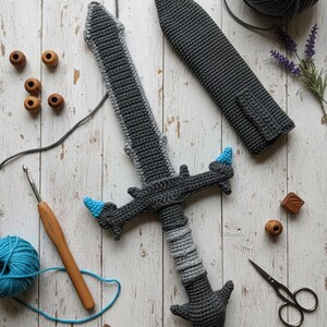 May include: A handmade crochet sword in shades of gray with blue accents. The sword is accompanied by a matching sheath. Other craft supplies, including yarn, a crochet hook, and wooden beads, are also visible.