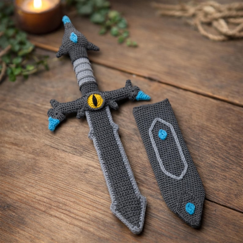 Crochet pattern for a fantasy sword and sheath, featuring a dark gray crocheted sword with a yellow eye detail on the hilt, turquoise accents, and a matching crocheted scabbard, displayed on a wooden surface.