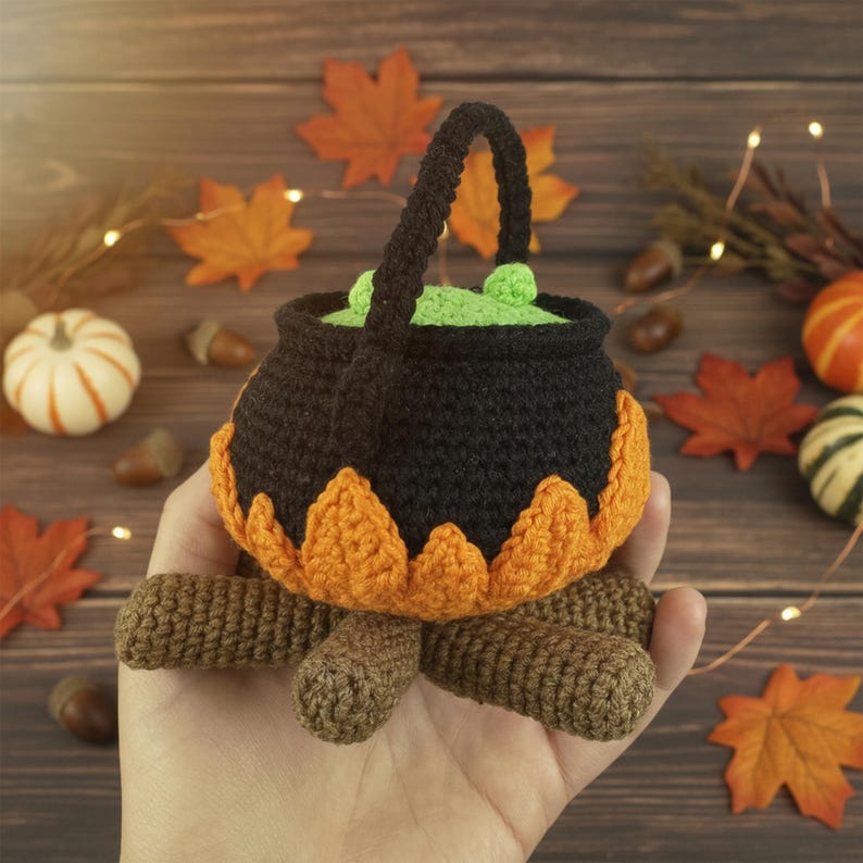 K&ouml;nnte beinhalten: Ein handgeh&auml;kelter Halloween-Kessel in Schwarz und Orange, gef&uuml;llt mit gr&uuml;nem Zaubertrank. Der Kessel ist von Herbstbl&auml;ttern, K&uuml;rbissen und Eicheln umgeben, was eine herbstliche Szene erzeugt.