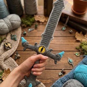 May include: Crocheted gray sword with a yellow and black eye design, and blue accents. The handmade toy sword is held in a hand, with yarn and other craft supplies in the background. A unique, whimsical item.