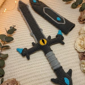 May include: Crocheted sword and sheath set in shades of gray, light gray, and blue. The sword features a yellow eye design and blue accents. The sheath is also gray with blue accents. The set is displayed on a cream-colored surface.