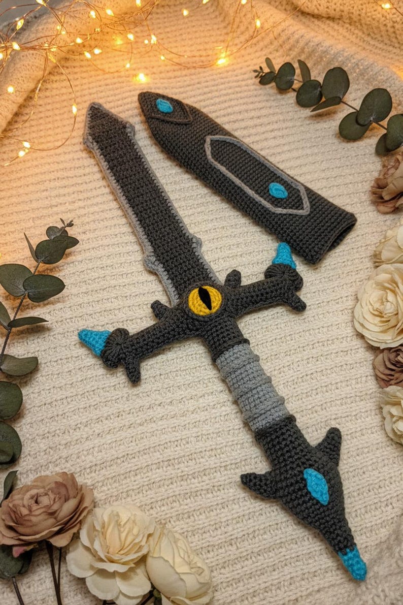 May include: Crocheted sword and sheath set in shades of gray, light gray, and blue. The sword features a yellow eye design and blue accents. The sheath is also gray with blue accents. The set is displayed on a cream-colored surface.